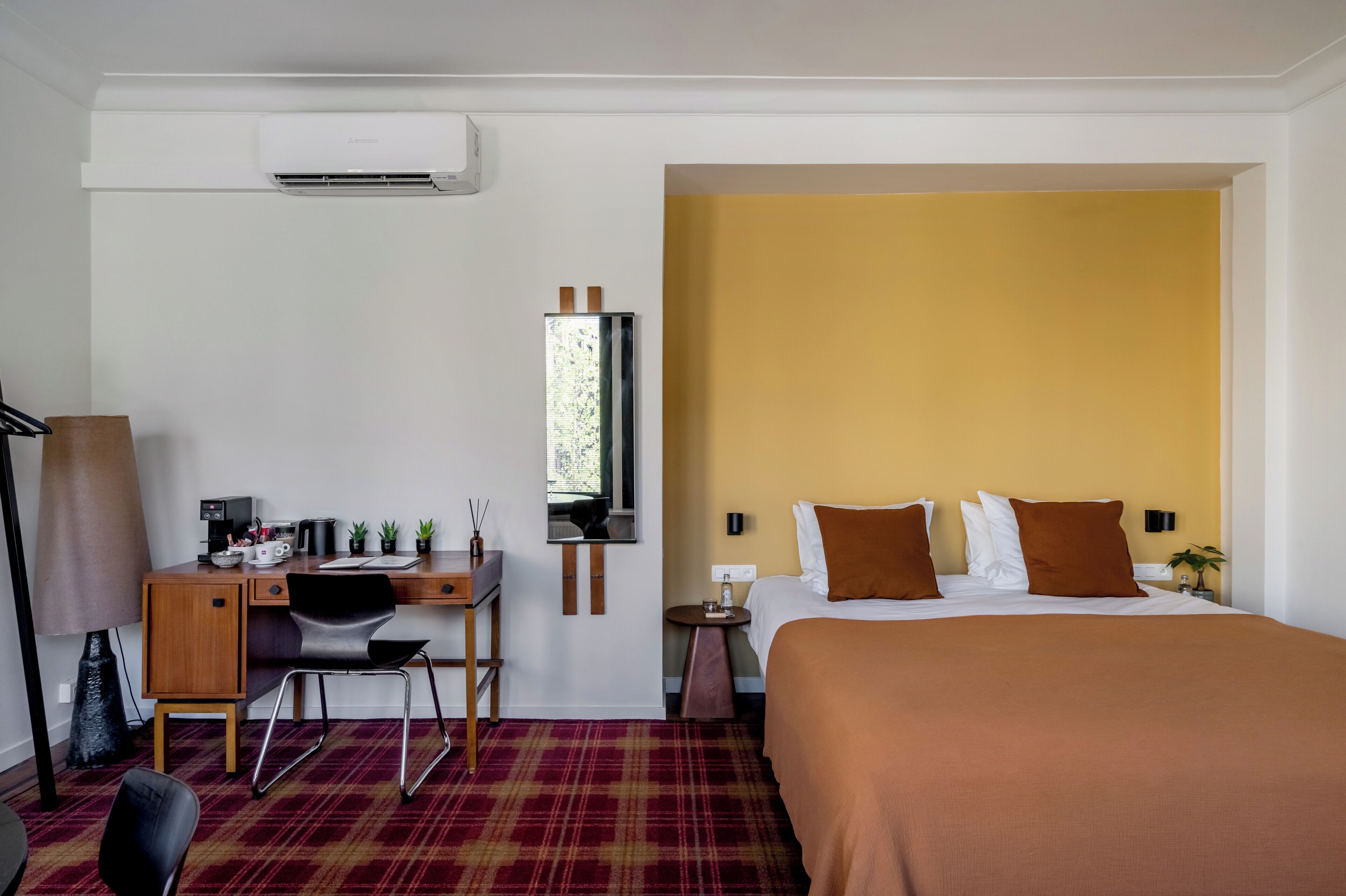 Una camera d'albergo con un letto, una scrivania, una sedia, una lampada e un condizionatore d'aria a parete.