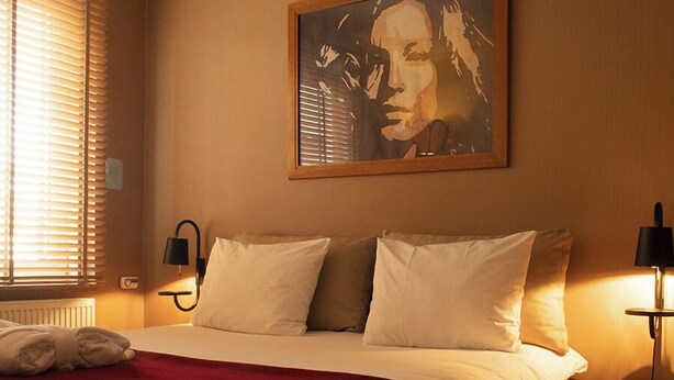 A hotel room with a bed, a painting on the wall, a window with blinds, and a bedside table with a lamp and books.
