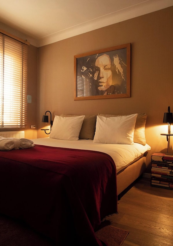 Ein Hotelzimmer mit Bett, einem Bild an der Wand, einem Fenster mit Jalousien sowie einem Nachttisch mit Lampe und Büchern.