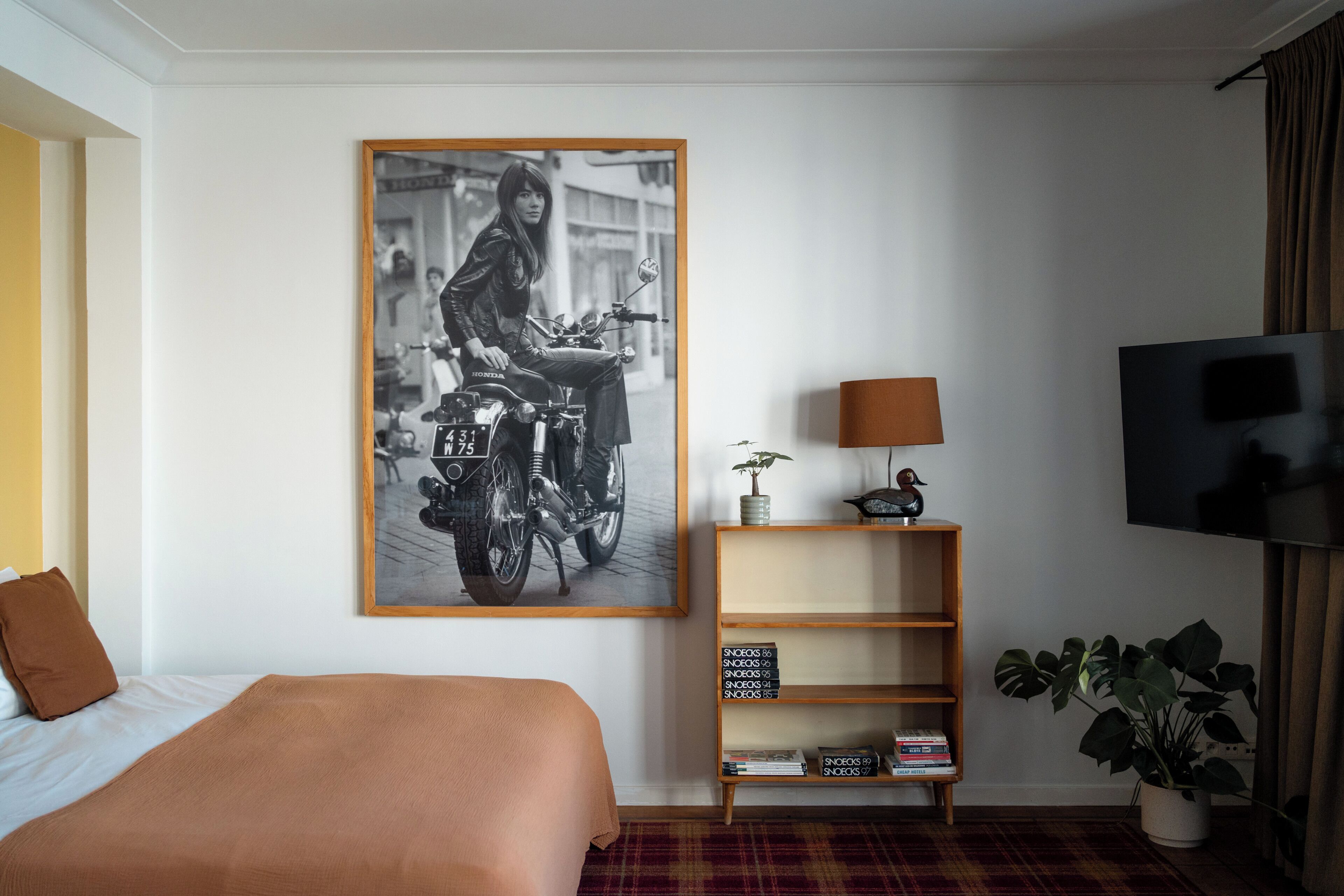 Ein Hotelzimmer mit einem Bett, einem gerahmten Foto einer Motorrad, einem Fernseher, einer Lampe, einem Regal mit Büchern und einer Pflanze.