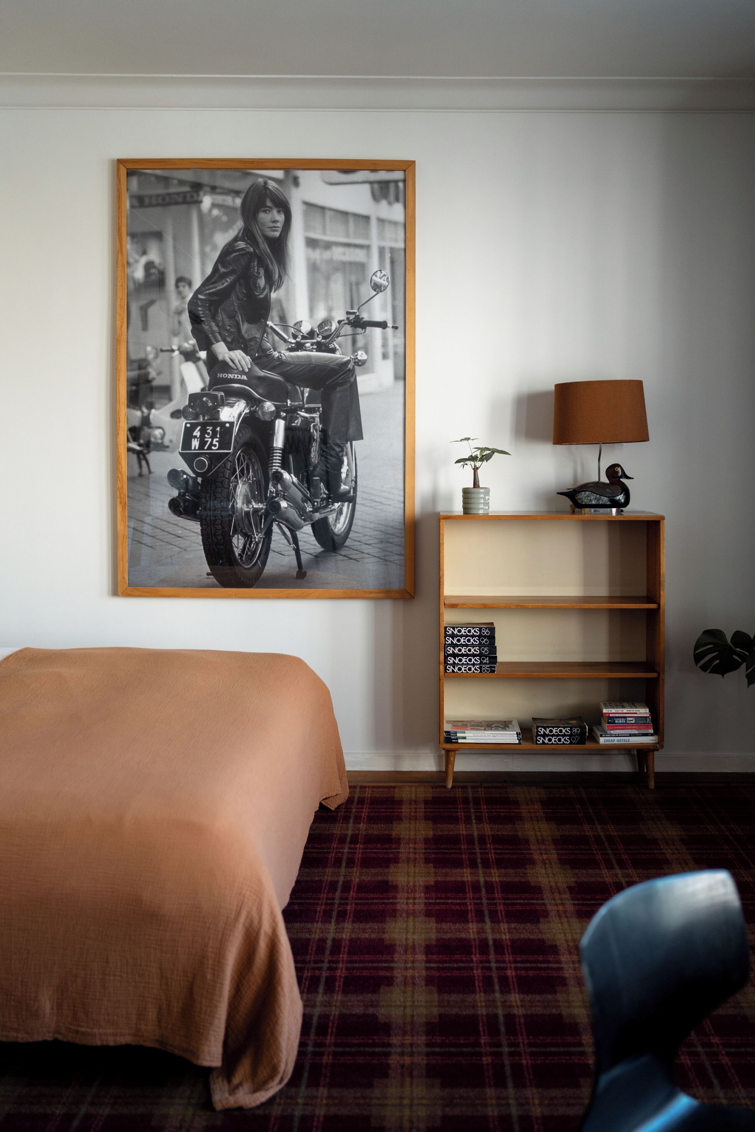 Une chambre d’hôtel avec un lit, une photo en noir et blanc encadrée représentant une moto accrochée au mur, une petite bibliothèque et une lampe.