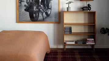Une chambre d’hôtel avec un lit, une photo en noir et blanc encadrée représentant une moto accrochée au mur, une petite bibliothèque et une lampe.