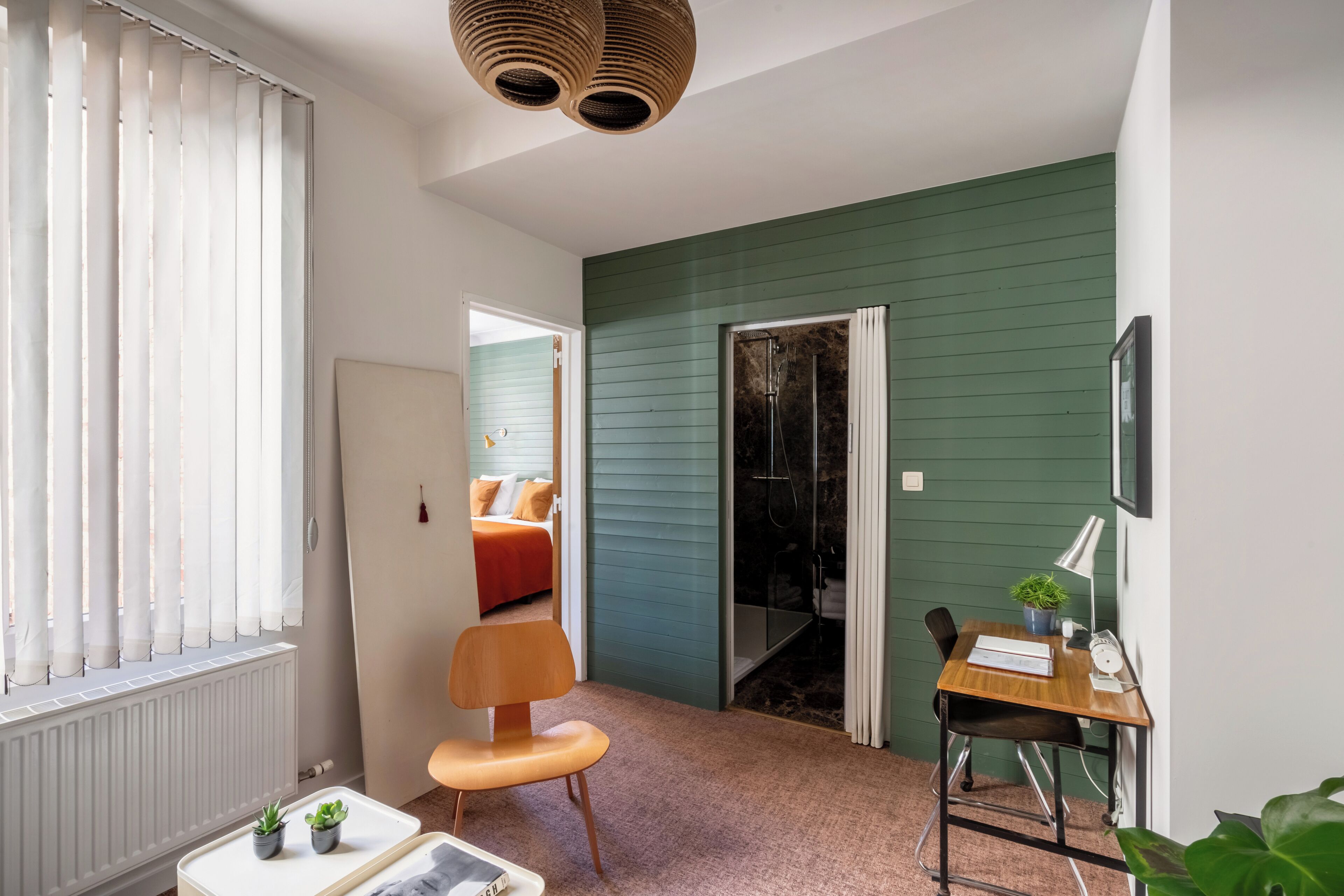 A compact room with a bed, a desk with a chair, a radiator, and a window with blinds.