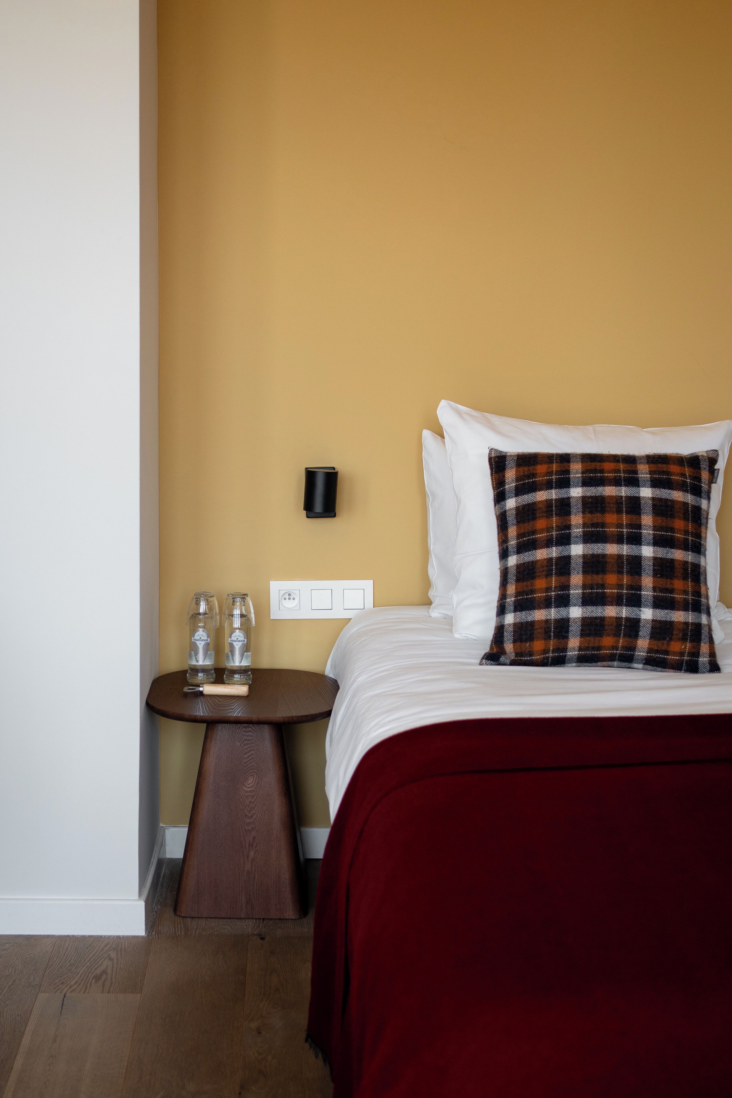 A hotel room with a bed, a red throw blanket, a plaid pillow, a small round table with two bottles, and a wall-mounted lamp.