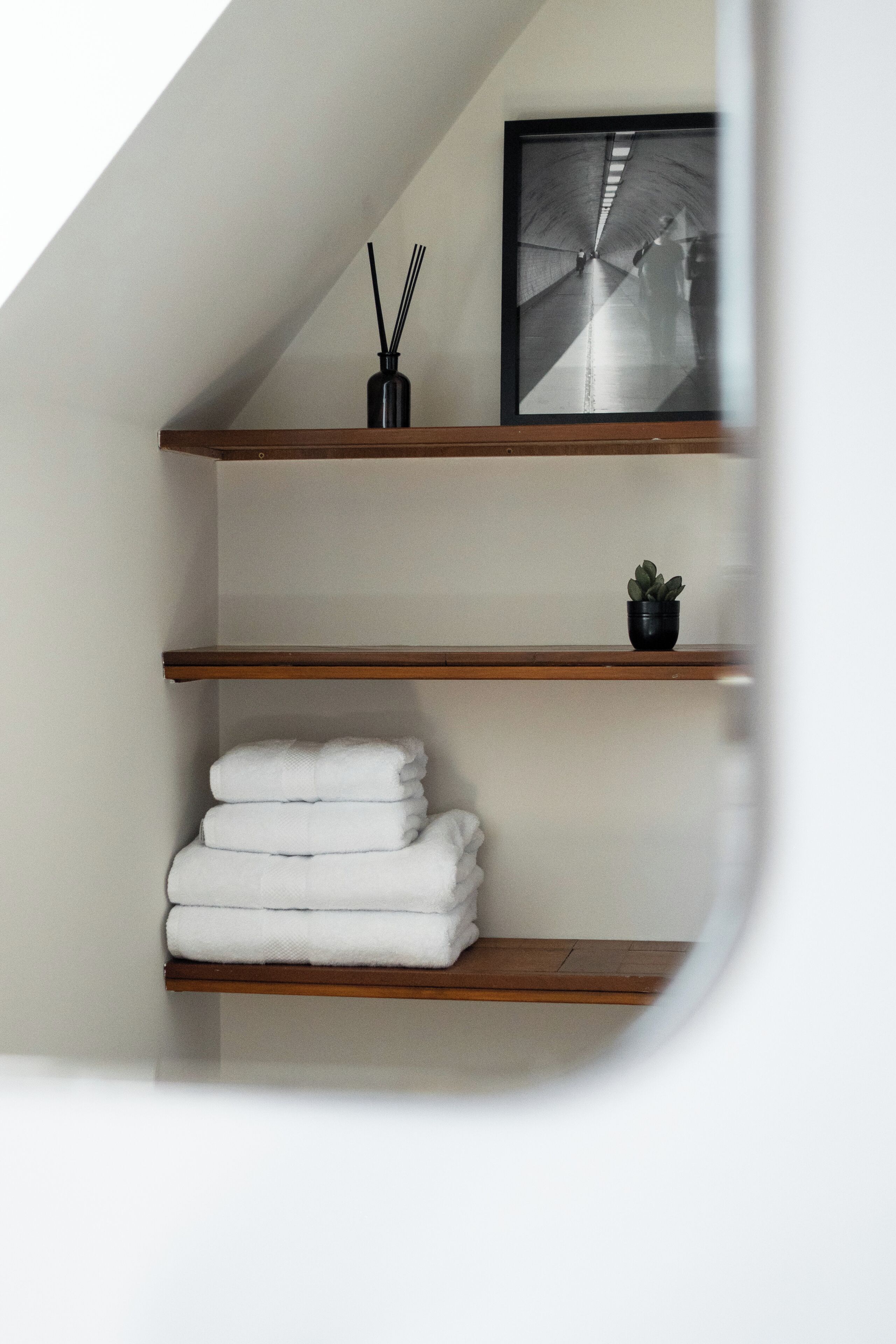 Una habitación con techo inclinado, estantes de madera, un cuadro enmarco, una pequeña planta en maceta y toallas blancas dobladas con cuidado.