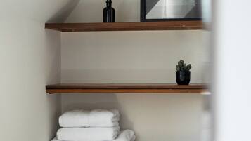 Una habitación con techo inclinado, estantes de madera, un cuadro enmarco, una pequeña planta en maceta y toallas blancas dobladas con cuidado.