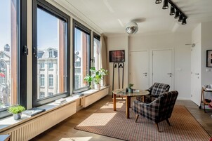 A modern living room with large windows, a round table, a plaid armchair, and a radiator.