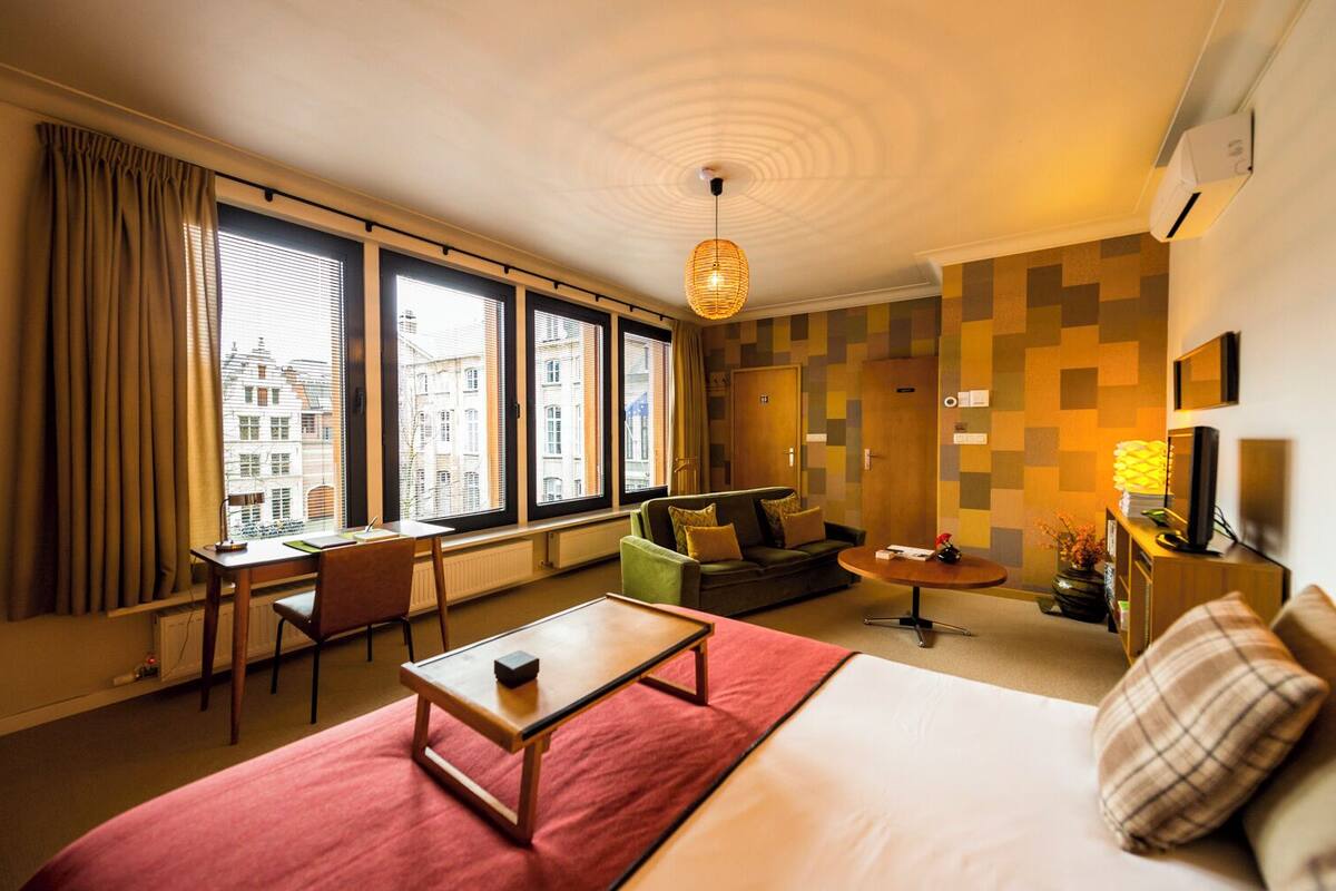 Ein Hotelzimmer mit einem großen Bett, einem Schreibtisch mit Stuhl, einer Couch, einem kleinen runden Tisch und einem Fernseher. Große Fenster mit Vorhängen, eine Wand-Klimaanlage und Blick auf Gebäude im Freien.