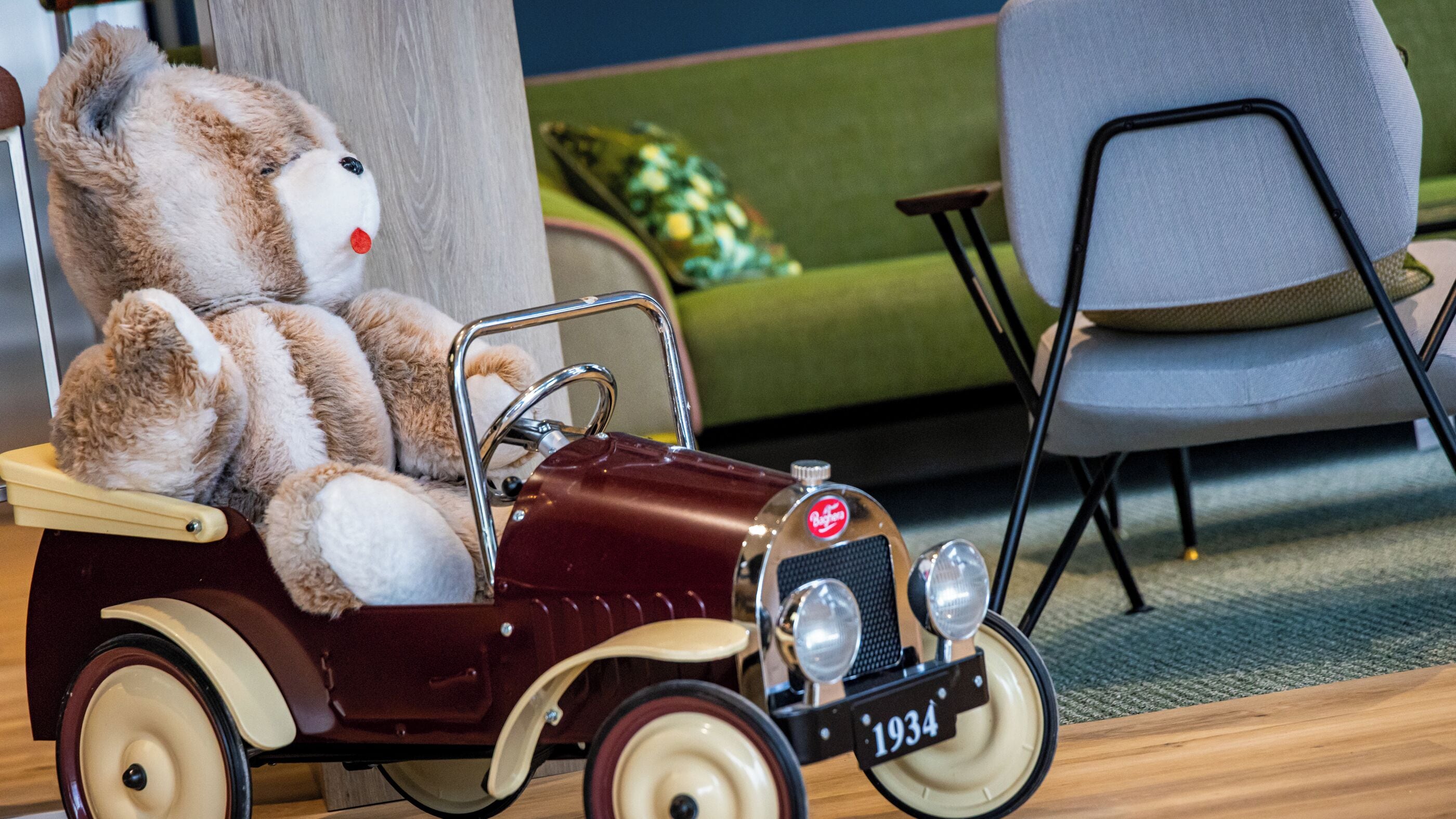 A vintage toy car with a teddy bear in the driver's seat, a vintage car license plate reading "1934", and a modern grey chair in the background.