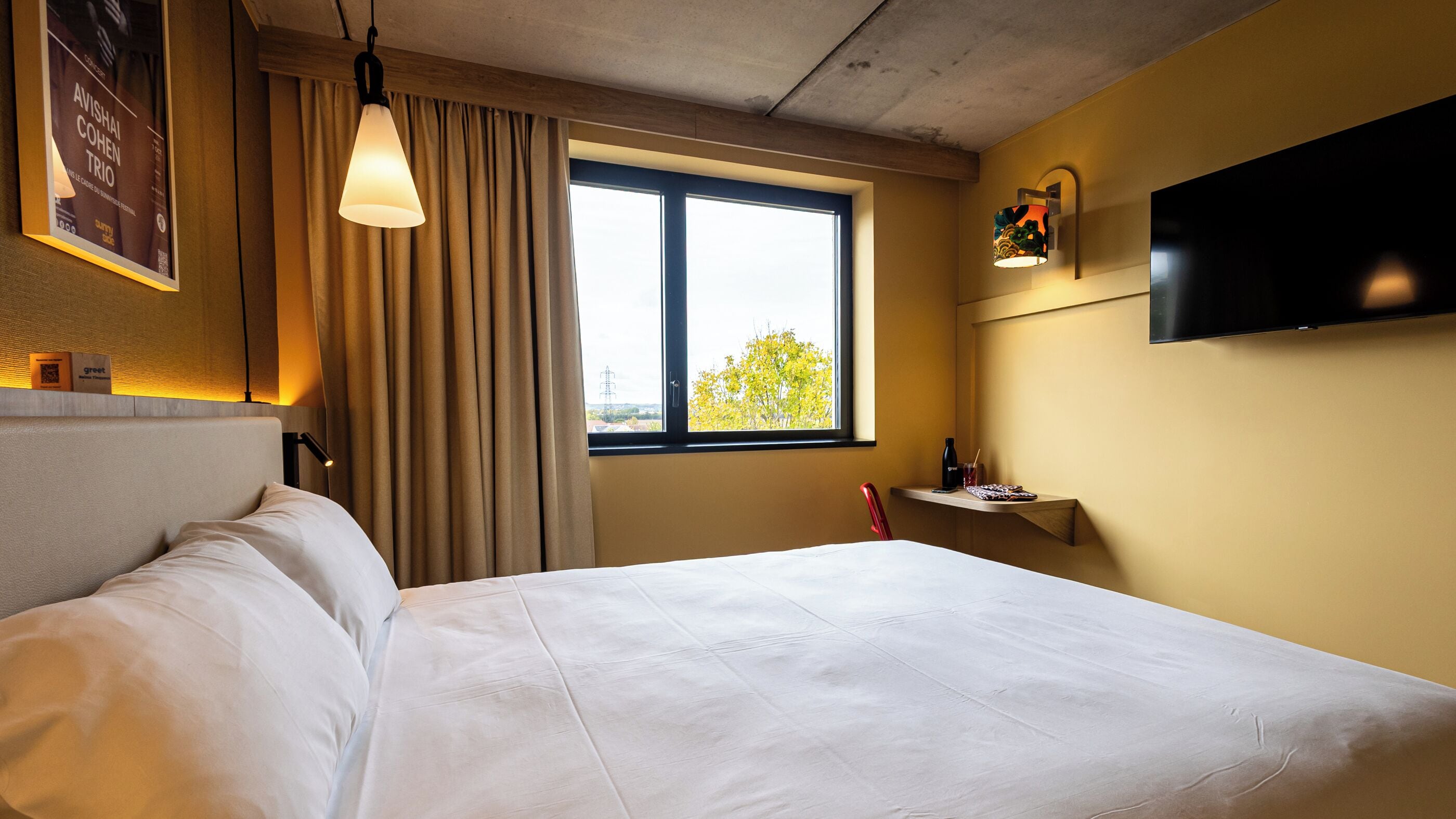 A hotel room with a bed, a window with curtains, a mounted TV, and a small shelf with items.