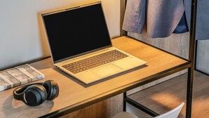 Una computadora portátil sobre un escritorio de madera, con audífonos, un libro y una chaqueta colgada en la pared.