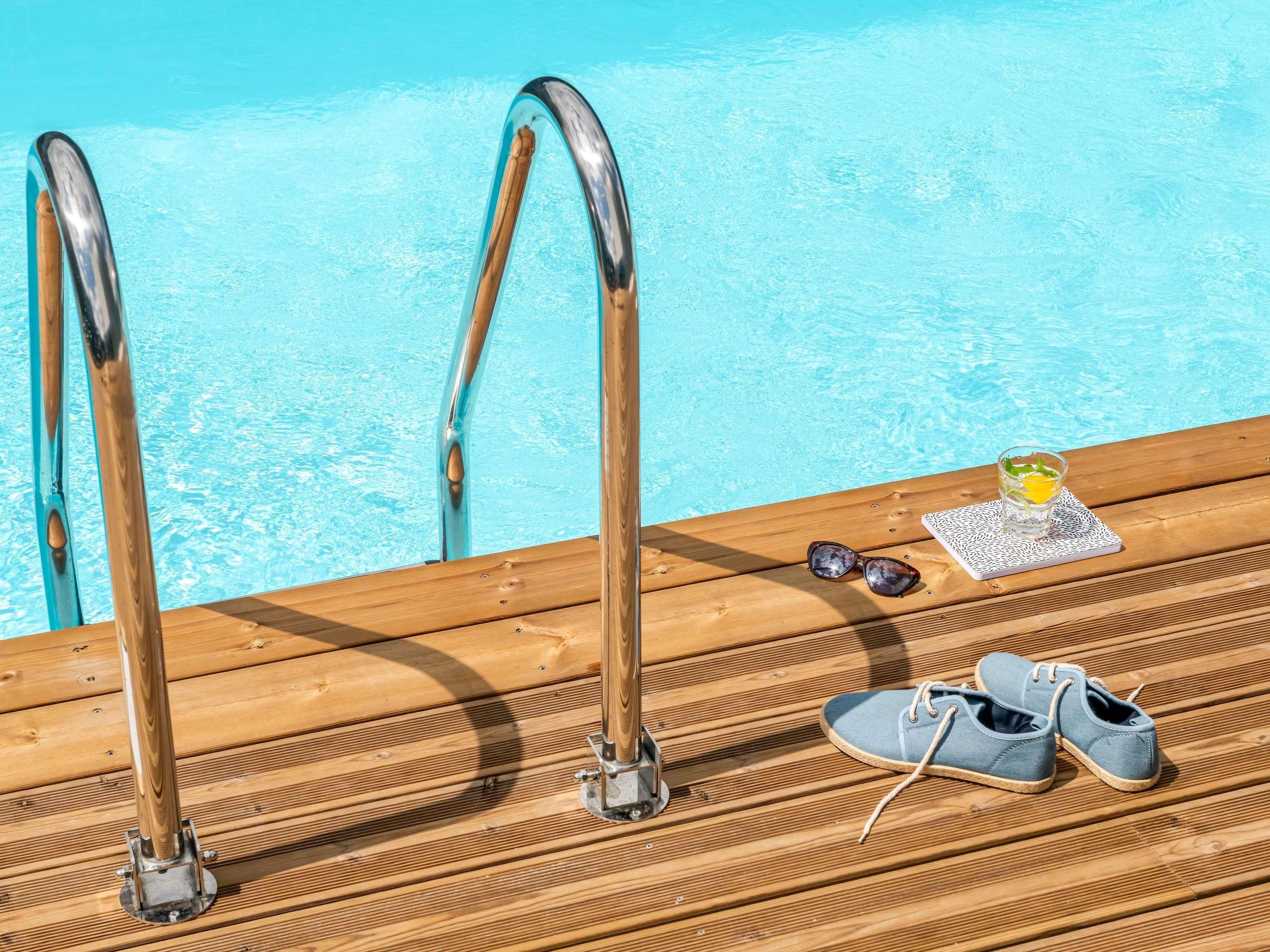 Terraza de madera con vistas a la piscina, un par de zapatos, gafas de sol y un vaso con una bebida.