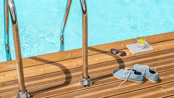 Terraza de madera con vistas a la piscina, un par de zapatos, gafas de sol y un vaso con una bebida.
