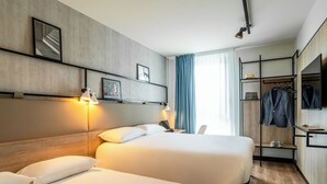 Una habitación de hotel con dos camas, un escritorio con lámpara, un perchero con ropa y un televisor en la pared.