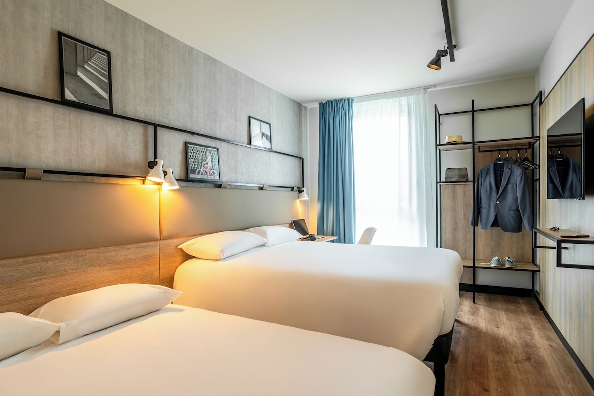 Una habitación de hotel con dos camas, un escritorio con lámpara, un perchero con ropa y un televisor en la pared.