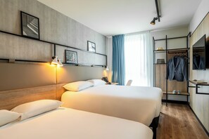 Une chambre d’hôtel avec deux lits, un bureau avec une lampe, un porte-manteau avec des vêtements et un téléviseur fixé au mur.