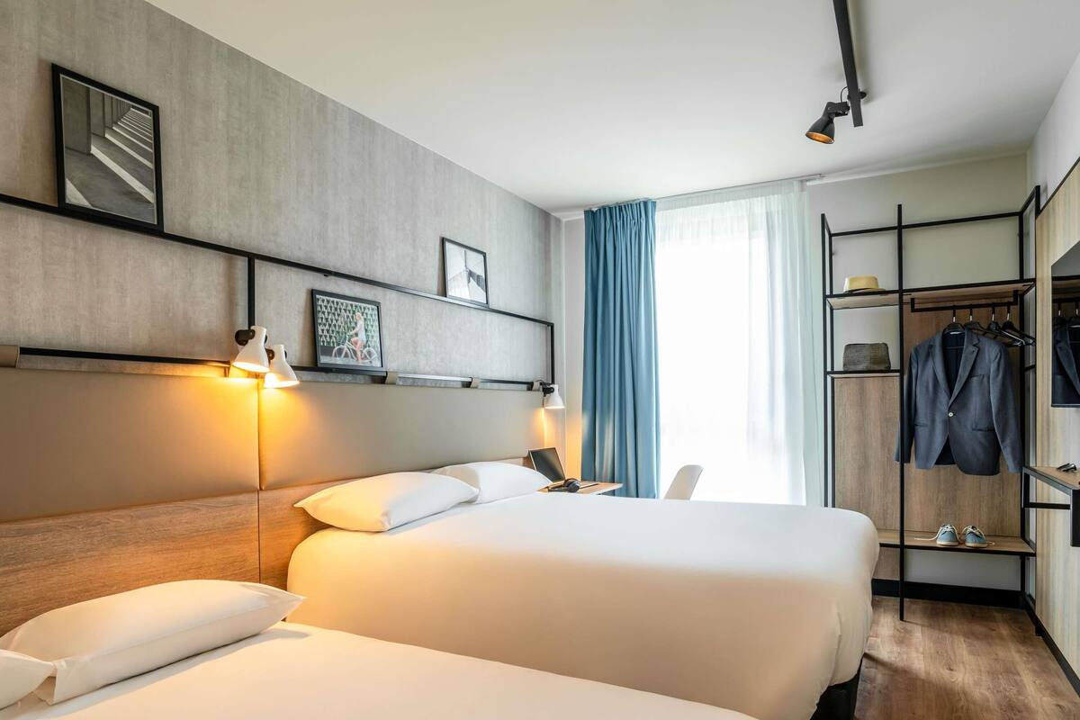 Ein Hotelzimmer mit zwei Betten, einem Schreibtisch mit Stuhl, einem Kleiderschrank mit Kleidung und einem großen Fenster mit Vorhängen.
