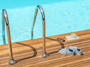 En træterrasse med udsigt til poolen, et par sko, solbriller og et glas med drik.