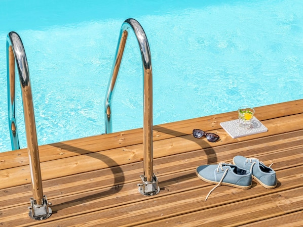 Eine Holzterrasse mit Blick auf ein Schwimmbecken, ein Schuhpaar, Sonnenbrille und ein Glas mit Getränk.