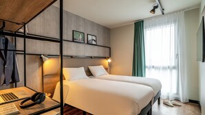 Habitación de hotel con dos camas, un escritorio con una computadora portátil y auriculares, y un estante de pared con una chaqueta y fotografías.