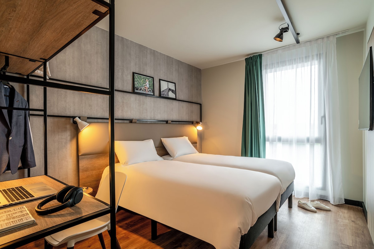 Habitación de hotel con dos camas, un escritorio con una computadora portátil y auriculares, y un estante de pared con una chaqueta y fotografías.