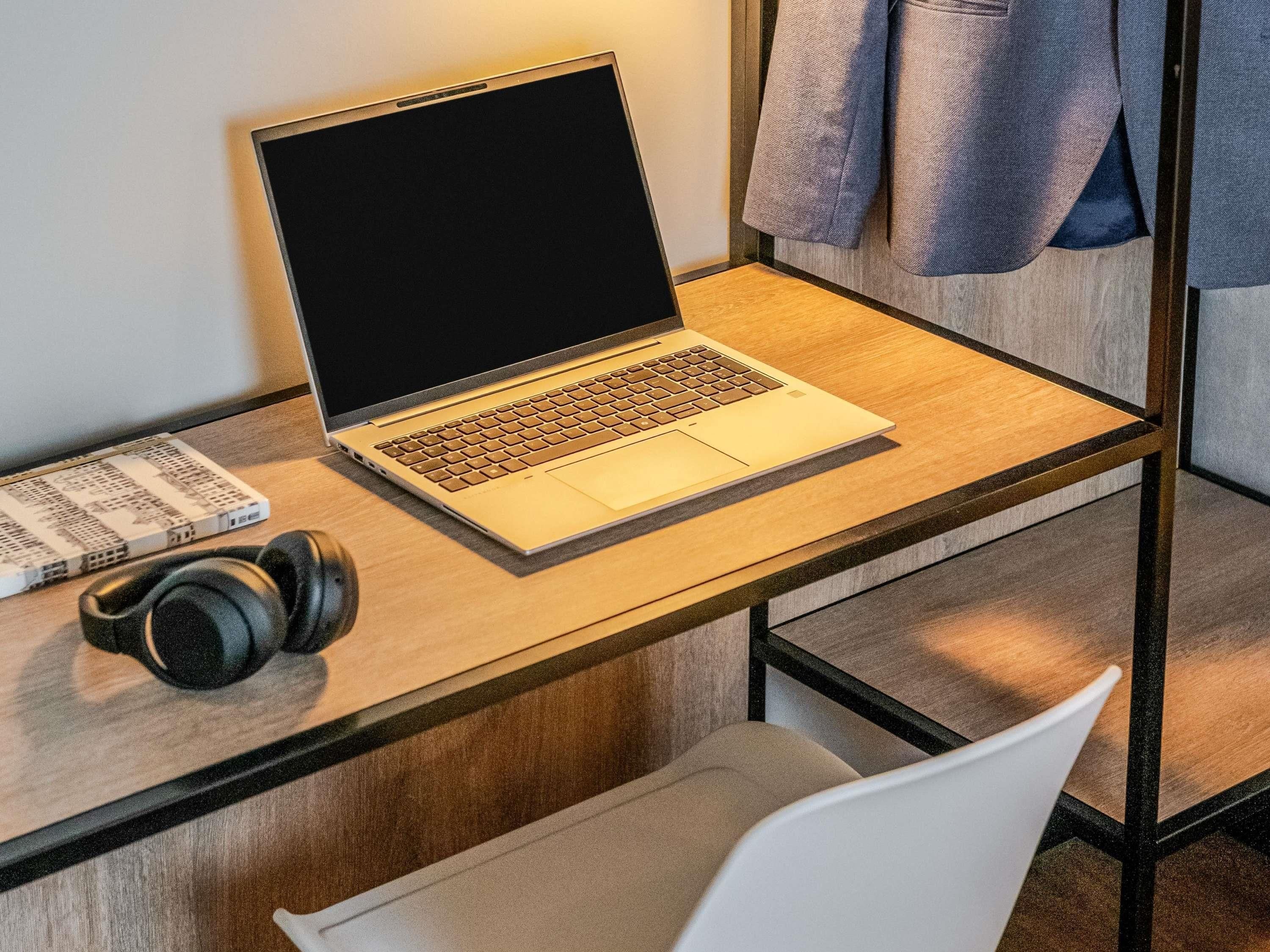 Una computadora portátil sobre un escritorio de madera, con auriculares, un libro y una chaqueta colgada en un perchero.