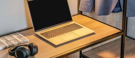 Una computadora portátil sobre un escritorio de madera, con auriculares, un libro y una chaqueta colgada en un perchero.