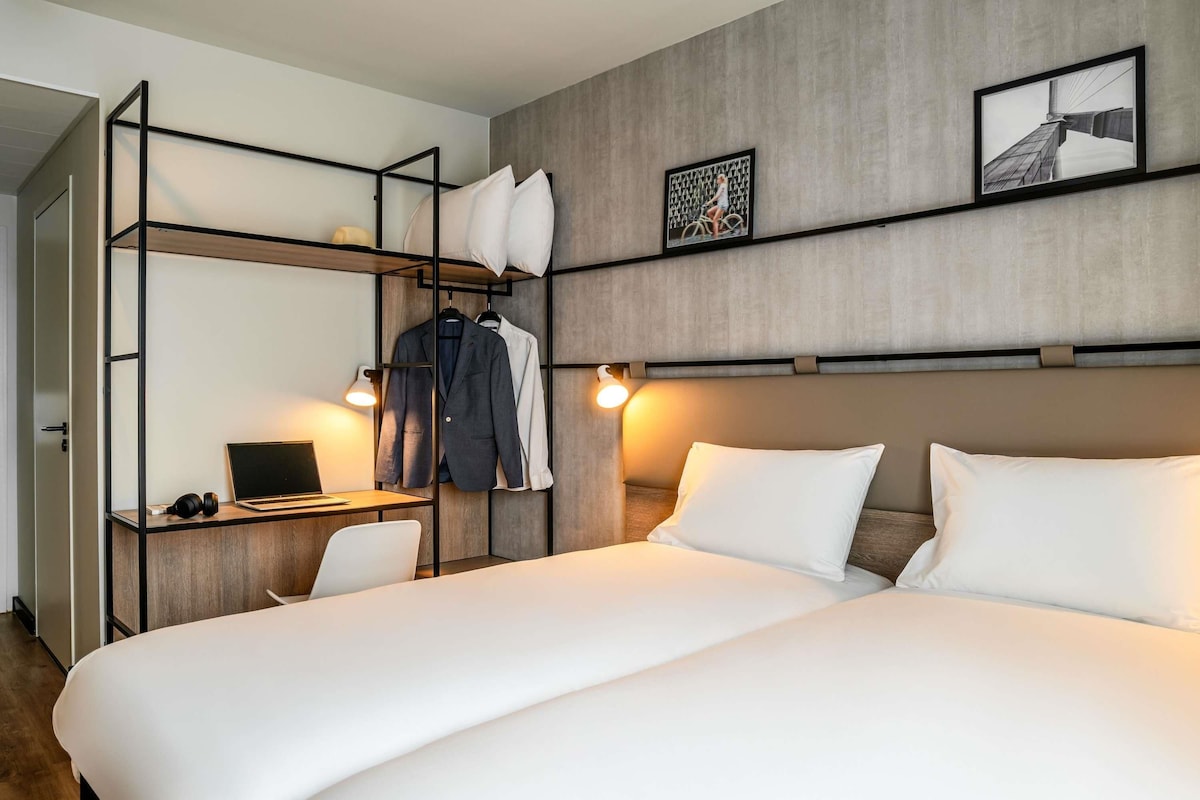 A hotel room with a bed, a desk with a laptop, a chair, a wardrobe, and two wall-mounted lights.