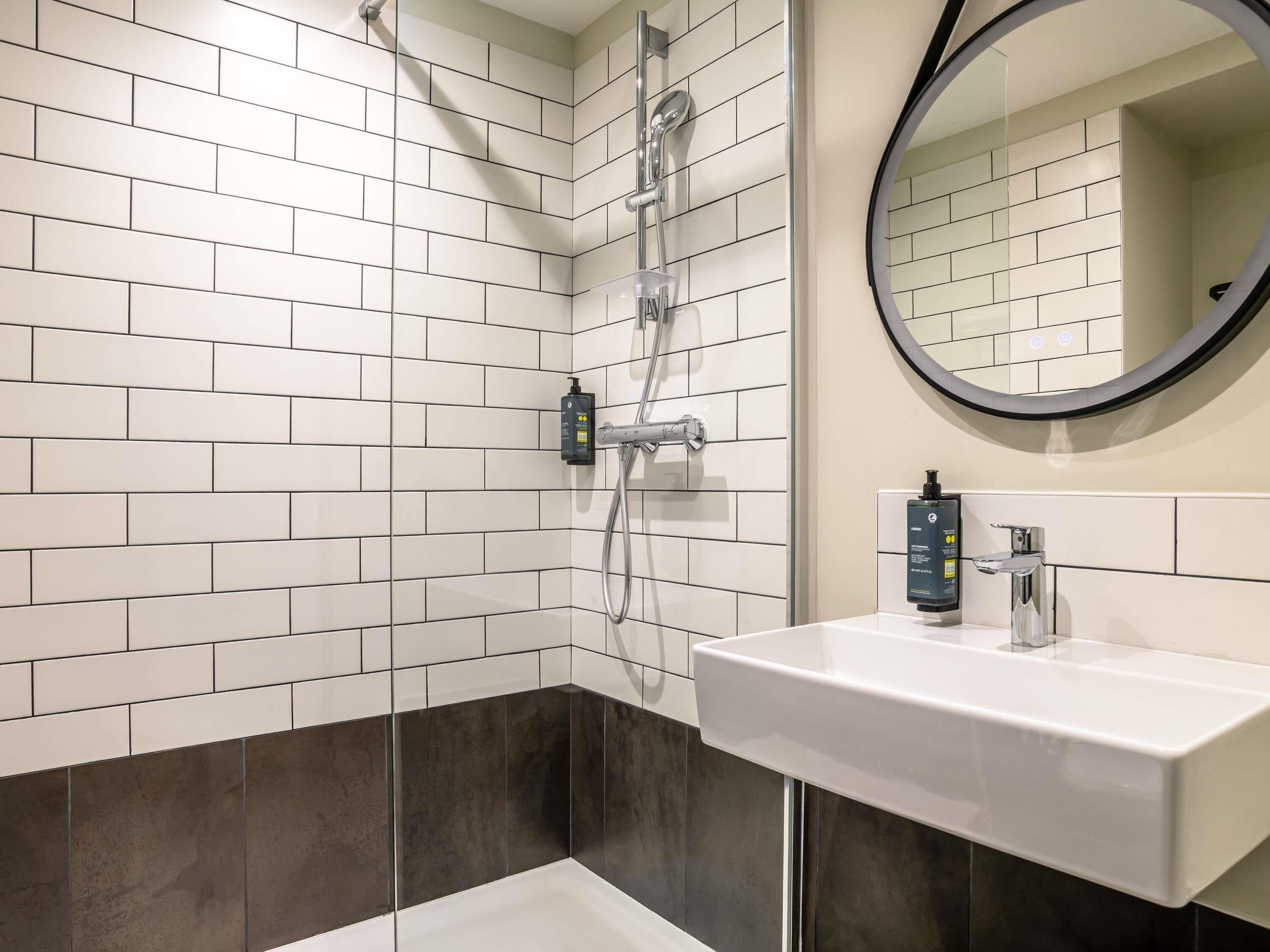A modern bathroom with white and dark tiles, a shower with a handheld showerhead, a round mirror, and a white sink with a faucet.
