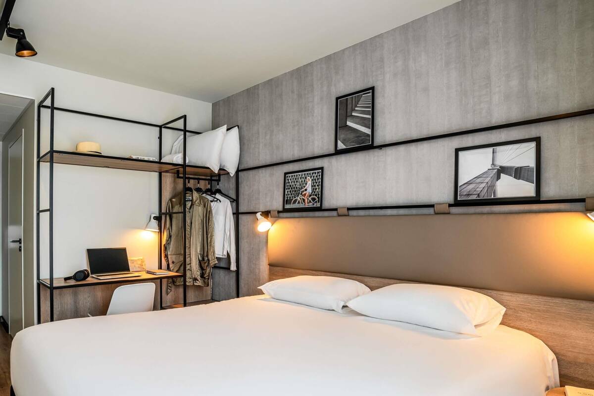 A modern hotel room with a large bed, a desk with a laptop, and a wardrobe with hanging clothes.