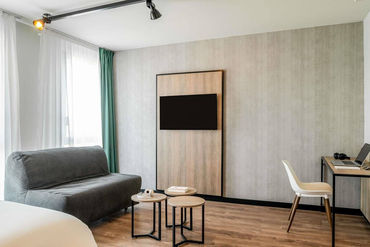 A modern hotel room with a grey sofa, a small round table with a pair of shoes, a desk with a laptop, and a mounted TV.