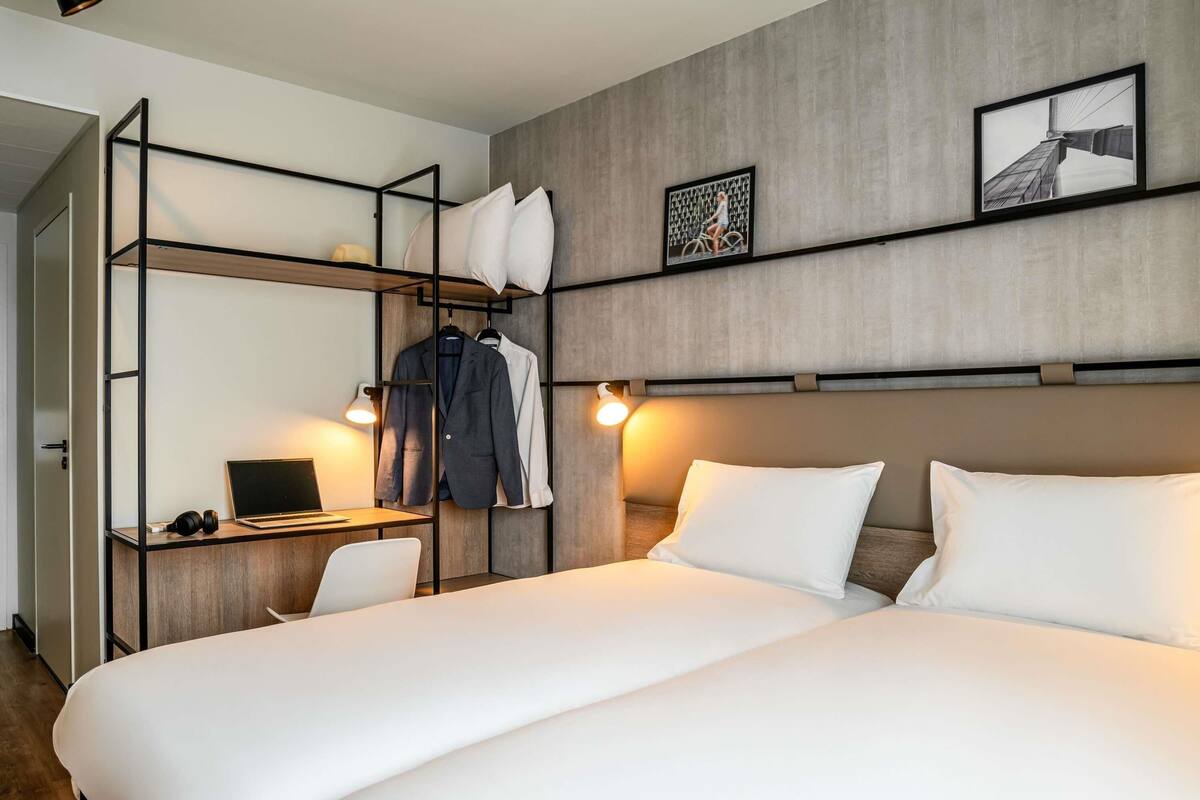 A hotel room with a bed, a desk with a laptop, a chair, a wardrobe, and two wall-mounted lights.