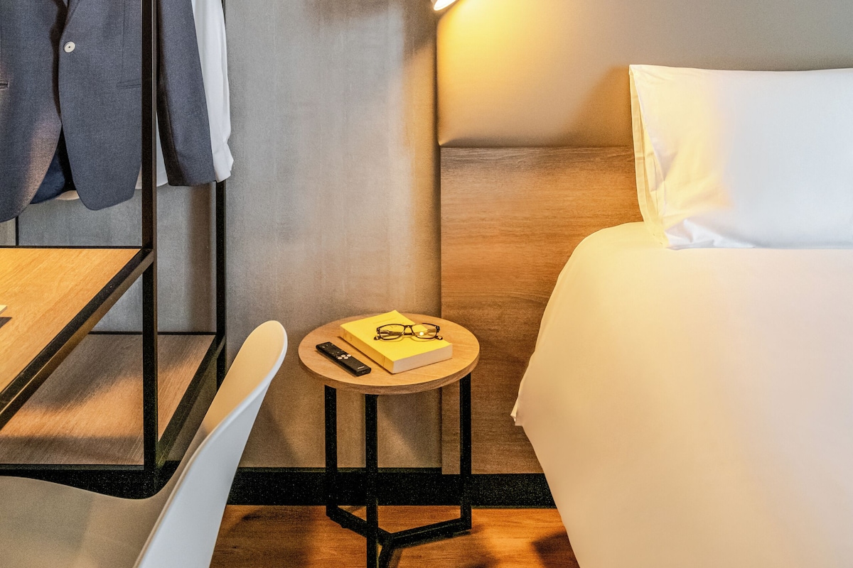 Habitación de hotel con literas, una mesita de noche con lámpara, un cuadro enmarcado en la pared y una pequeña mesa redonda con un libro y gafas.