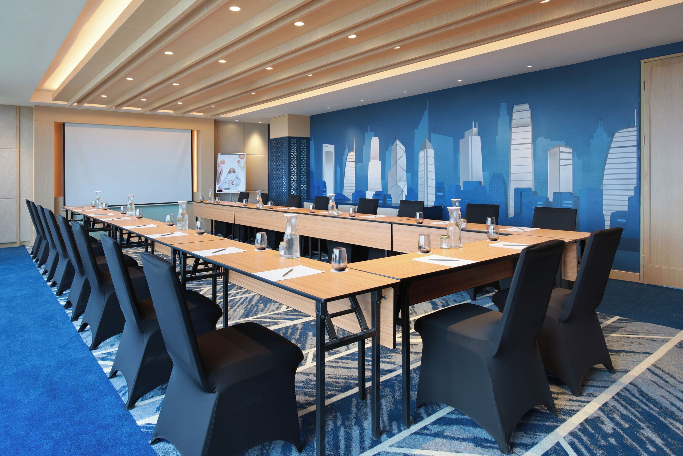 A conference room with a long table, chairs, a projector screen, and a cityscape mural on the wall.