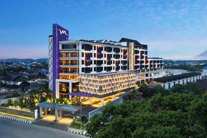 Un hotel de varios pisos con un llamativo logo 'M', una entrada cubierta y una zona ajardinada al frente.