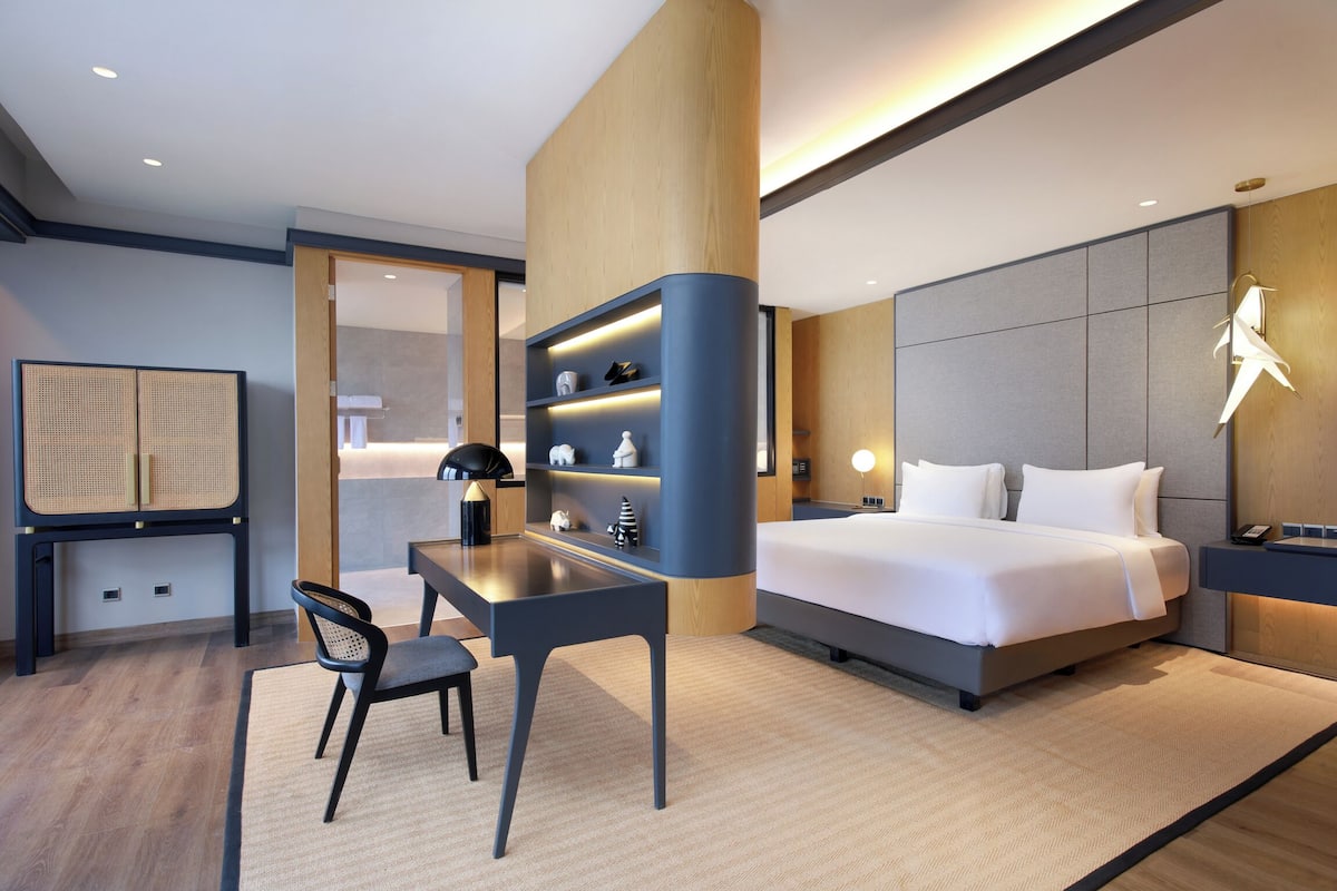 A modern hotel room with a bed, desk, chair, and shelves.