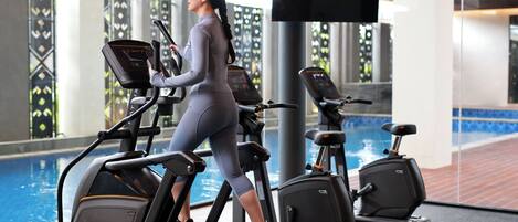 A woman using an elliptical machine in a gym with a pool in the background.