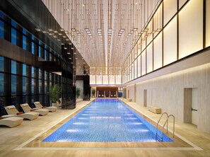 Une piscine intérieure moderne, de forme longue et étroite, entourée d’espaces de détente avec des chaises longues et des plantes en pot.