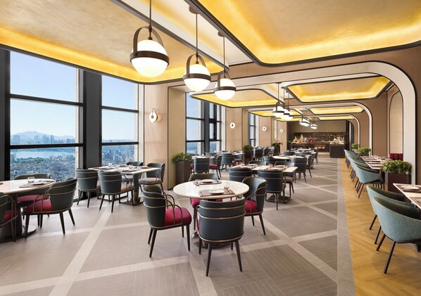Un vaste espace dédié à la salle à manger, avec des tables et des chaises rondes, de grandes fenêtres offrant une vue sur la ville et un éclairage moderne à suspension.
