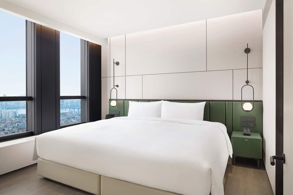 Ein modernes Hotelzimmer mit großem Bett, Blick auf die Stadt, grünem Kopfteil und Nachttischen mit Lampen.