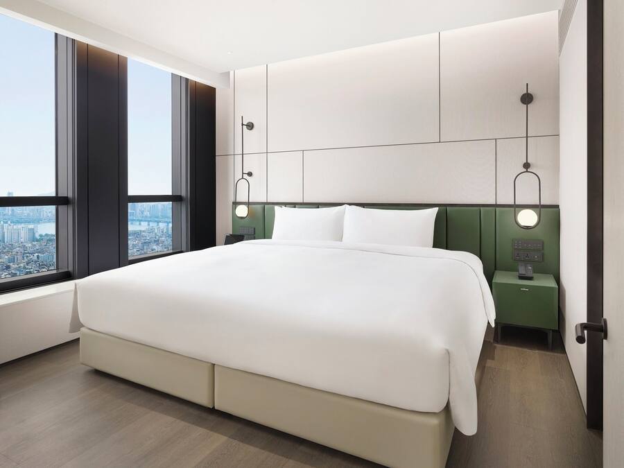 Habitación de hotel moderna con una cama grande, vista a la ciudad, cabecera verde y mesitas de noche con lámparas.