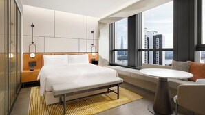 Una habitación de hotel moderna con una cama grande, un banco, una mesa redonda y vistas de la ciudad a través de amplias ventanas.