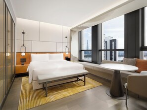 Une chambre d’hôtel moderne avec un grand lit, un banc, une table ronde et une vue sur le paysage urbain grâce à de grandes fenêtres.