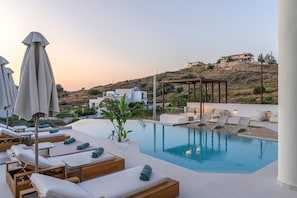 Área à beira da piscina com espreguiçadeiras e guarda-sóis, vista desobstruída de um morro, e um edifício moderno ao fundo.
