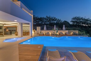 Uma área de piscina moderna com espreguiçadeiras e guarda-sóis, rodeada por um jardim ajardinado e um edifício com fachada de vidro.