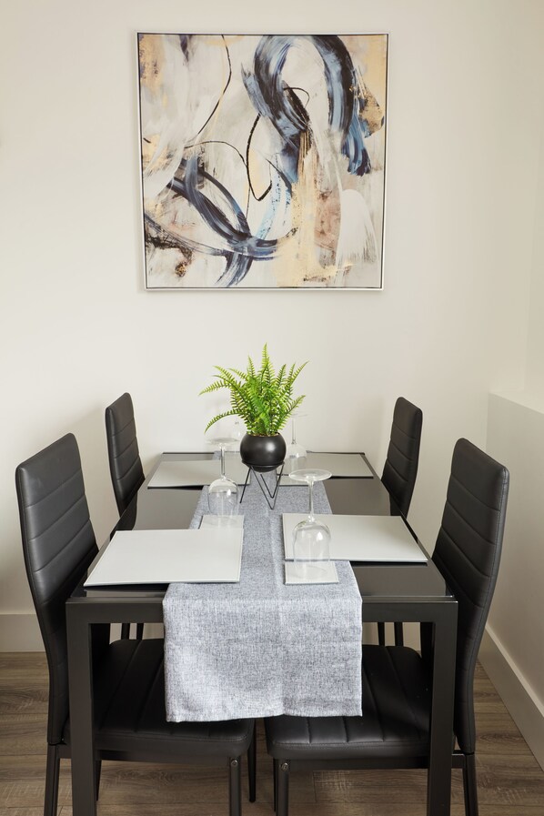 A dining table with a modern abstract painting above it, black chairs, a potted plant, and a grey table runner.