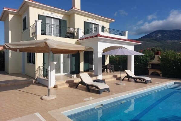A two-story house with a swimming pool, outdoor furniture, and a mountain backdrop. - Two bedroomed villa, each with balcony and ensuite, a private pool, and BBQ. (Karavados)