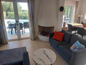 A living room with a fireplace, a sofa, a coffee table, and a view of an outdoor pool area. - Two bedroomed villa, each with balcony and ensuite, a private pool, and BBQ. (Karavados)