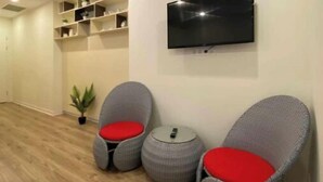Una stanza moderna con una TV a schermo piatto montata a parete, due poltrone grigie in vimini con cuscini rossi, un tavolino rotondo e una pianta in vaso.
