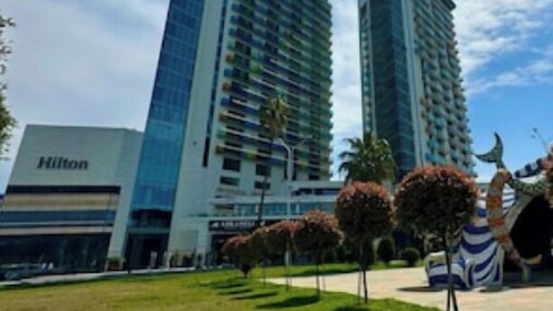 Un hotel moderno con facciata in vetro, cartello Hilton ben visibile e un prato curato con alberi.